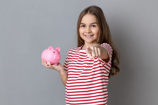 Portrait of cute positive little girl wearing striped T-shirt standing pointing at camera, holding piggybank, saving money, banking. Indoor studio shot isolated on gray background.
