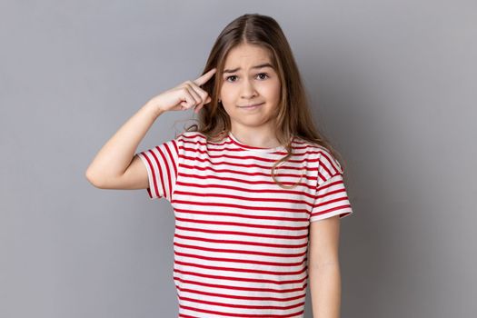 Are you kidding me. Portrait of little girl wearing striped T-shirt holding finger near head temple and gesturing, looking displeased with crazy idea. Indoor studio shot isolated on gray background.