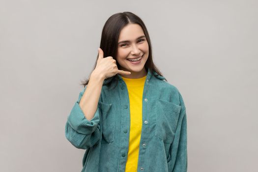 Dark haired woman making telephone gesture near head and smiling playfully, waiting for call, wants to talk on phone, wearing casual style jacket. Indoor studio shot isolated on gray background.