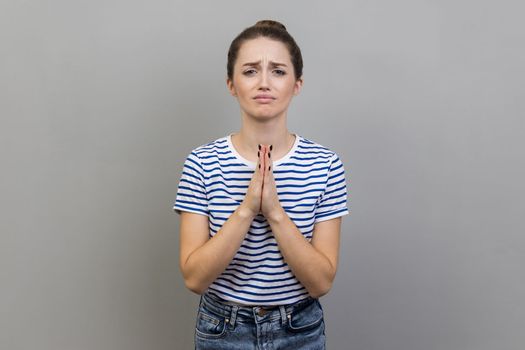 Please, I need. Portrait of woman wearing striped T-shirt holding hands in prayer and asking for help with pleading imploring expression, full of hope. Indoor studio shot isolated on gray background.
