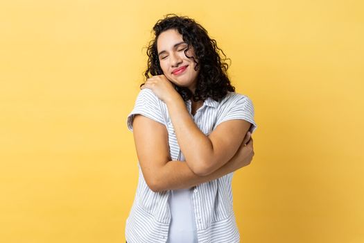 Portrait of delighted woman with dark wavy hair hugging herself and smiling with dreamy satisfied expression, love myself gesture. Indoor studio shot isolated on yellow background.