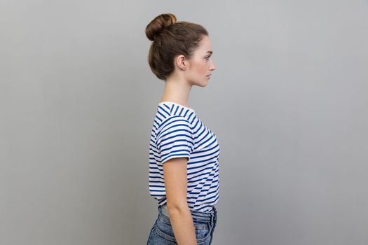 Side view portrait of beautiful serious bossy woman with bun hairstyle wearing striped T-shirt standing looking ahead with strict facial expression. Indoor studio shot isolated on gray background.