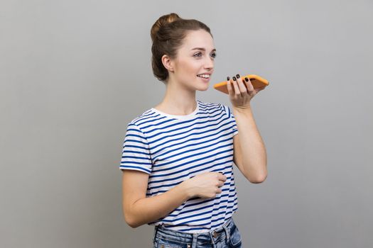 Smart voice technology. Portrait of woman wearing striped T-shirt talking to cell phone using virtual assistant, digital speaker application. Indoor studio shot isolated on gray background.
