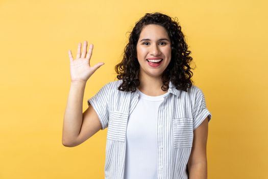 Portrait of friendly positive beautiful woman with dark wavy hair smiles toothily, raises palm greets friend, saying hello or goodbye. Indoor studio shot isolated on yellow background.
