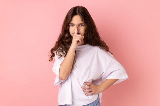 Portrait of serious concentrated little girl wearing white T-shirt holding finger on lips, making hush silence gesture, asking to keep secret. Indoor studio shot isolated on pink background.