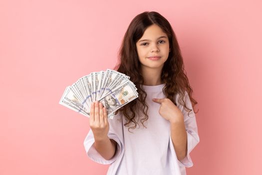 Portrait of adorable cute little girl wearing white T-shirt pointing at dollars banknotes in her hand, having calm expression, big profit. Indoor studio shot isolated on pink background.