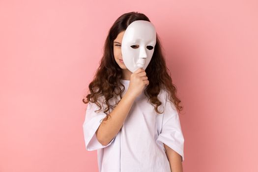 Portrait of dark haired little girl wearing white T-shirt covering face, standing with frowning face, multiple personality disorder. Indoor studio shot isolated on pink background.