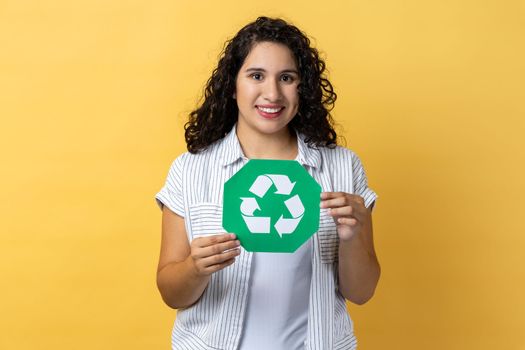 Portrait of attractive delighted woman with dark wavy hair holding green recycling sign, calls on to sort rubbish, save our planet from pollution. Indoor studio shot isolated on yellow background.