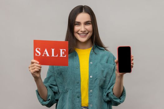 Cheerful woman holding sale inscription, cellphone and smiling at camera, showing discounts on mobile device, application, wearing casual style jacket. Indoor studio shot isolated on gray background.