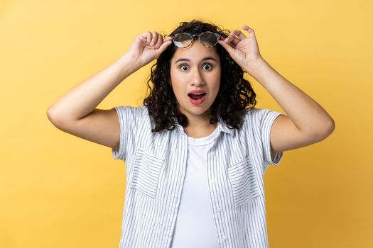 Portrait of surprised astonished woman with dark wavy hair standing raised eyeglasses and looking at camera with big eyes, sees something shocked. Indoor studio shot isolated on yellow background.