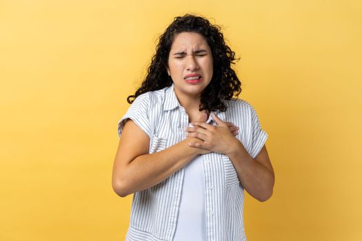 Portrait of sick attractive woman with dark wavy hair holding hand on chest, feeling awful acute pain in heart, risk of heart attack. Indoor studio shot isolated on yellow background.