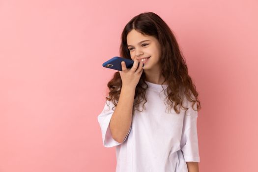 Mobile smart voice technology. Portrait of little girl wearing white T-shirt talking to smartphone using virtual assistant, digital speaker app. Indoor studio shot isolated on pink background.