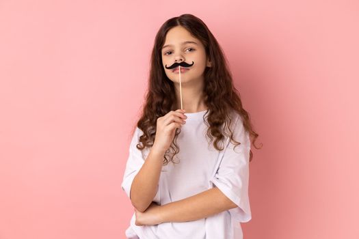 Portrait of little girl wearing white T-shirt keeping fake paper mustache on lips and looking at camera with serious humorous expression. Indoor studio shot isolated on pink background.