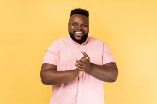 Portrait of positive friendly man wearing pink shirt standing with palms together, applauding, clapping hands congratulating you with victory. Indoor studio shot isolated on yellow background.