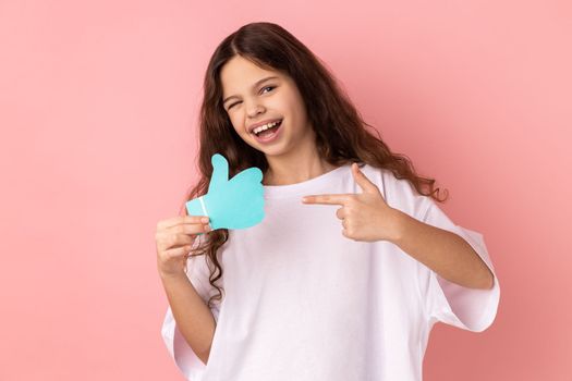 Portrait of happy satisfied adorable little girl wearing white T-shirt holding and pointing at like or thumbs up paper shape sign. Indoor studio shot isolated on pink background.