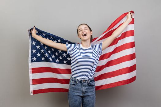 Portrait of cheerful young adult attractive woman wearing striped T-shirt holding big american flag, expressing happiness, screaming happily, rejoicing. Indoor studio shot isolated on gray background.