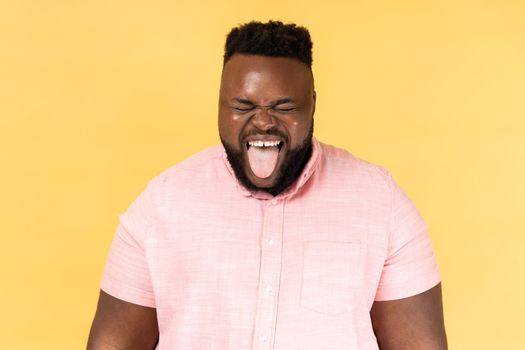 Portrait of man wearing pink shirt sticking out tongue and looking at camera, teasing with naughty expression, disobedient behavior. Indoor studio shot isolated on yellow background.