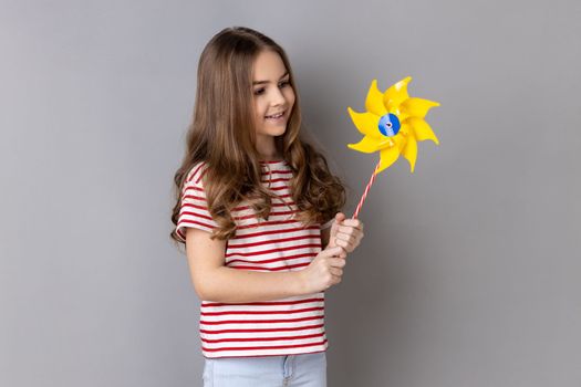 Portrait of positive optimistic little girl wearing striped T-shirt smiling carefree to camera, holding paper windmill, pinwheel toy on stick. Indoor studio shot isolated on gray background.