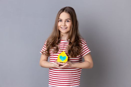 Portrait of little girl wearing striped T-shirt holding in hands little paper house looking at camera with smile, dreaming about apartment. Indoor studio shot isolated on gray background.