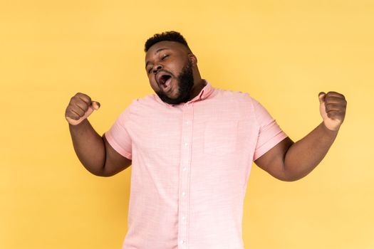 Portrait of tired bearded man wearing pink shirt yawning, feeling exhausted, lack of sleep, outstretching arms, keeps eyes closed. Indoor studio shot isolated on yellow background.