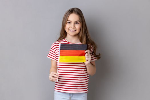 Germany flag. Portrait of satisfied optimistic adorable cute little girl wearing striped T-shirt holding german flag, looking at camera. Indoor studio shot isolated on gray background.
