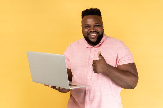 Portrait of smiling man holding laptop in hand showing thumbs up gesture, blogger likes posts, looking at camera, recommending to followers. Indoor studio shot isolated on yellow background.