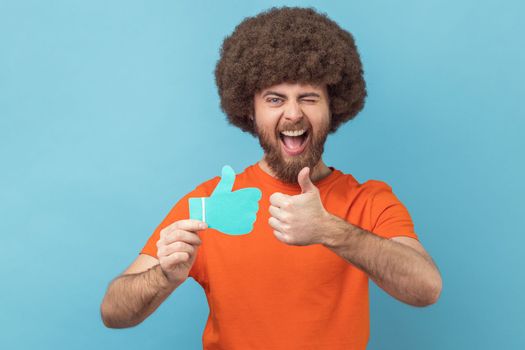 Man with Afro hairstyle wearing orange T-shirt holding and showing like or thumbs up paper shape sign, looking winking at camera with toothy smile. Indoor studio shot isolated on blue background.