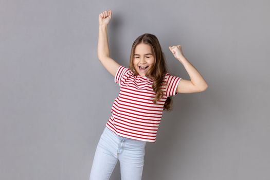 Portrait of joyful little girl wearing striped T-shirt expressing winning gesture with raised fists and screaming, celebrating victory. Indoor studio shot isolated on gray background.