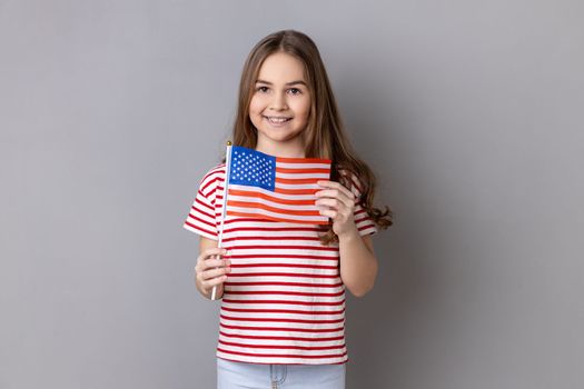 American flag. Portrait of delighted happy adorable cute little girl wearing striped T-shirt holding flag of United States of America, looking at camera. Indoor studio shot isolated on gray background