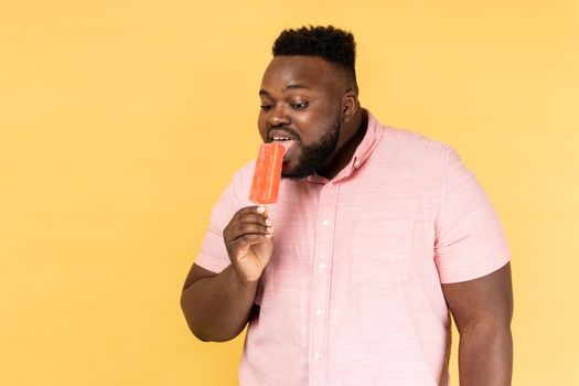 Portrait of positive optimistic man wearing pink shirt holding licking sweet ice cream, trying delicious confectionery dessert. Indoor studio shot isolated on yellow background.
