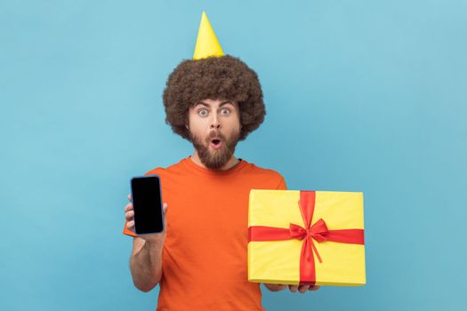 Shocked man with Afro hairstyle wearing orange T-shirt and party cone holding gift box and mobile phone with blank display for promotion. Indoor studio shot isolated on blue background.
