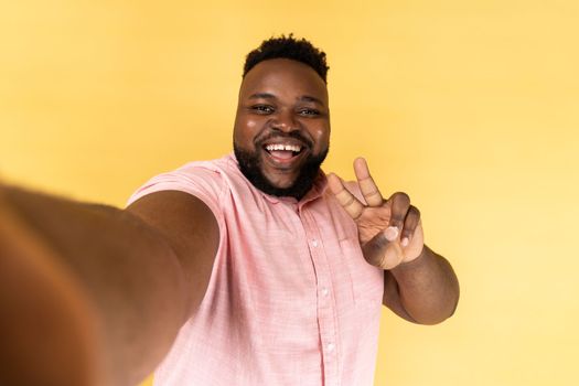 Portrait of happy man wearing pink shirt taking selfie, looking at camera POV, point of view of photo, showing victory gesture. Indoor studio shot isolated on yellow background.