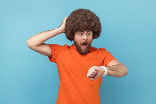 Portrait of man with Afro hairstyle wearing orange T-shirt pointing finger on his wrist watch with nervous face, time is out, keeps hand on head. Indoor studio shot isolated on blue background.