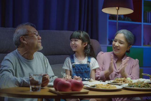 Happy Asian family grandmother grandfather and granddaughter dining on table and having fun during at home night time, senior parent and child eating food together in living room indoors together