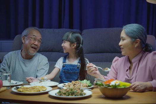 Happy Asian senior parent and child eating dinner food together in living room indoors, family grandmother grandfather and granddaughter dining on table and having fun during at home night time