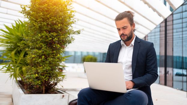 Business on the Go Businessman Working on his Laptop on the Street near office building. Handsome hispanic business person sitting use laptop. Confident male manager working outdoor.