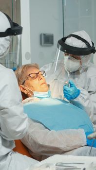 Woman at dentist while treatment process in coronavirus pandemic lying on stomatological chair. Doctor and nurse working wearing coverall, face shield, protection suit, mask, gloves in dental office