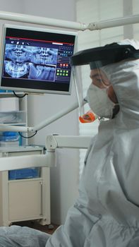 Dentist doctor with face shield pointing on stomatological monitor explaining dental x-ray to patient during global pandemic. Nurse and orthodontist wearing protection suit, coverall, mask and gloves