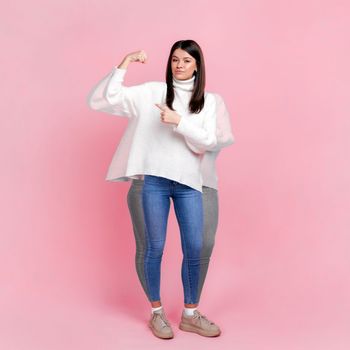 Full length portrait of happy woman before and after loosing her weight, pointing at biceps, lose weight through exercise and nutrition. Indoor studio shot isolated on pink background