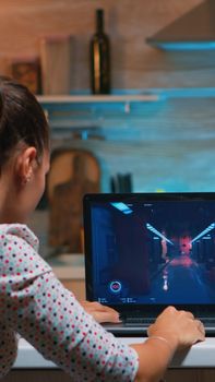 Woman gamer sitting at home in kitchen testing new game on professional laptop. Tired player gaming online video games on her personal computer with modern technology network wireless