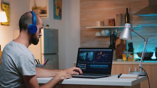 Professional videographer working in video editing app wearing headphones in front of laptop sitting in home kitchen. Freelancer processing audio film montage on professional laptop in midnight