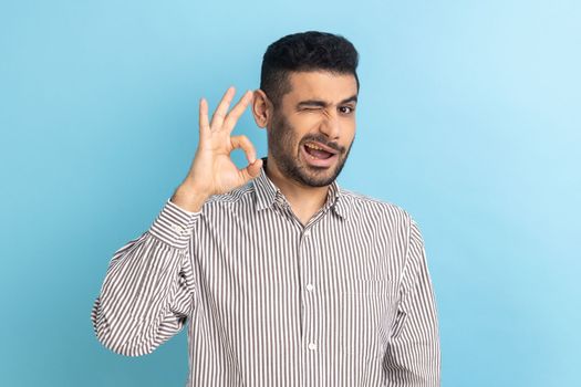 Portrait of delighted bearded businessman standing, looking at camera showing Ok sign gesture, expressing positive emotions, wearing striped shirt. Indoor studio shot isolated on blue background.