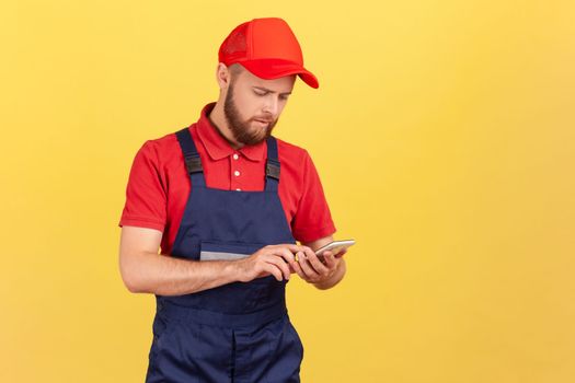 Serious handyman in overalls typing on mobile phone, using cellphone messenger to accept online order as delivery, repair and maintenance services. Indoor studio shot isolated on yellow background.