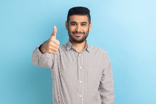 Portrait of businessman with beard standing with thumbs up, like gesture, demonstrating approval and agree with suggestion, wearing striped shirt. Indoor studio shot isolated on blue background.