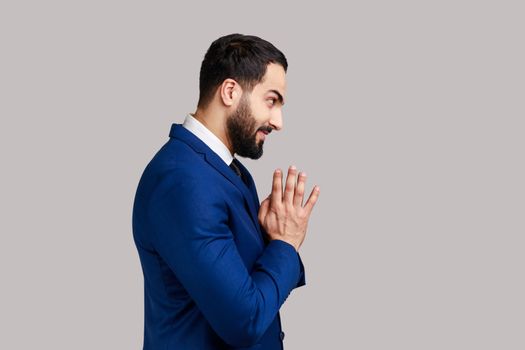 Side view of cunning bearded man clasping hands and planning evil tricky prank or scheming, cheating with sly smile, wearing official style suit. Indoor studio shot isolated on gray background.