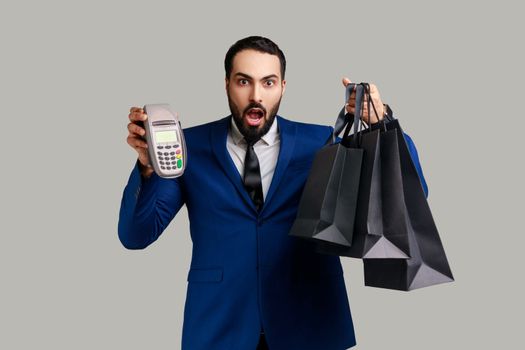 Surprised businessman holding payment terminal and paper shopping bags, easy express order and delivery, wearing official style suit. Indoor studio shot isolated on gray background.
