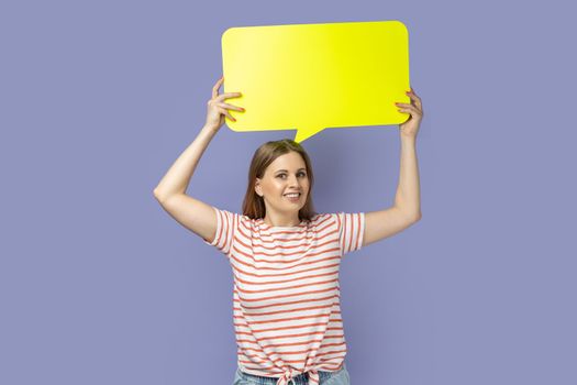 Portrait of positive smiling satisfied blond woman wearing striped T-shirt holding yellow bubble for text, looking at camera with toothy smile. Indoor studio shot isolated on purple background.