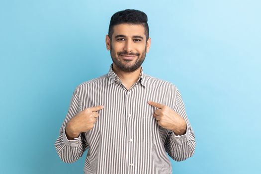 It's me. Portrait of ambitious bearded businessman with toothy smile, pointing fingers on himself, being confident, wearing striped shirt. Indoor studio shot isolated on blue background.