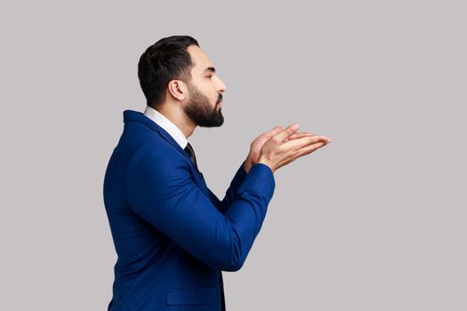 Side view of bearded man sending air kiss over palms, flirting to camera, demonstrating romantic amorous feelings, wearing official style suit. Indoor studio shot isolated on gray background.