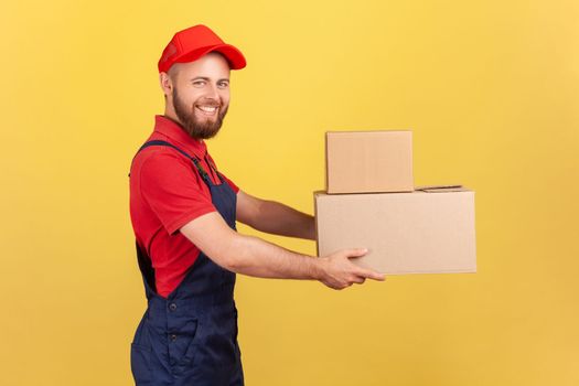 Side view of happy deliveryman holding two cardboard boxes, delivering order door-to-door, shipment and cargo transportation service. Indoor studio shot isolated on yellow background.
